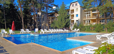 Basen zewnętrzny otoczony białymi leżakami i leżankami, położony na słonecznym dziedzińcu z zielonymi drzewami i nowoczesnym budynkiem mieszkalnym w tle, co tworzy relaksującą i przyjazną atmosferę