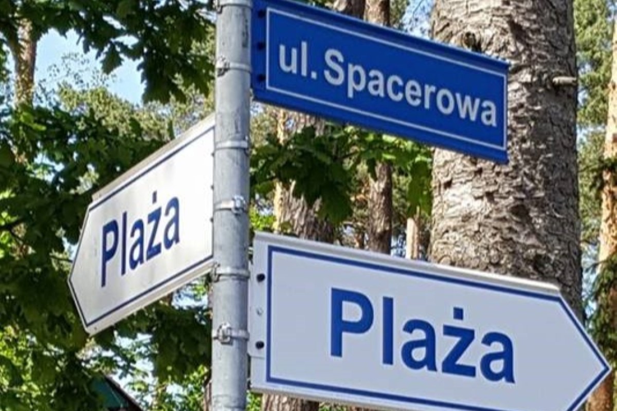 Trzy niebiesko-białe znaki drogowe umieszczone na słupie wśród wysokich drzew i zielonych liści. Górny znak wskazuje ulicę Spacerową, a dwa dolne znaki wskazują różne kierunki i oznaczają Plażę. Scena jest jasna i letnia, przywodząc na myśl przyjazną i relaksującą atmosferę w pobliżu plaży.